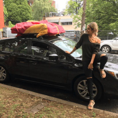 Michelle sitting on the hood of her black car, which has camping gear strapped to the roof
